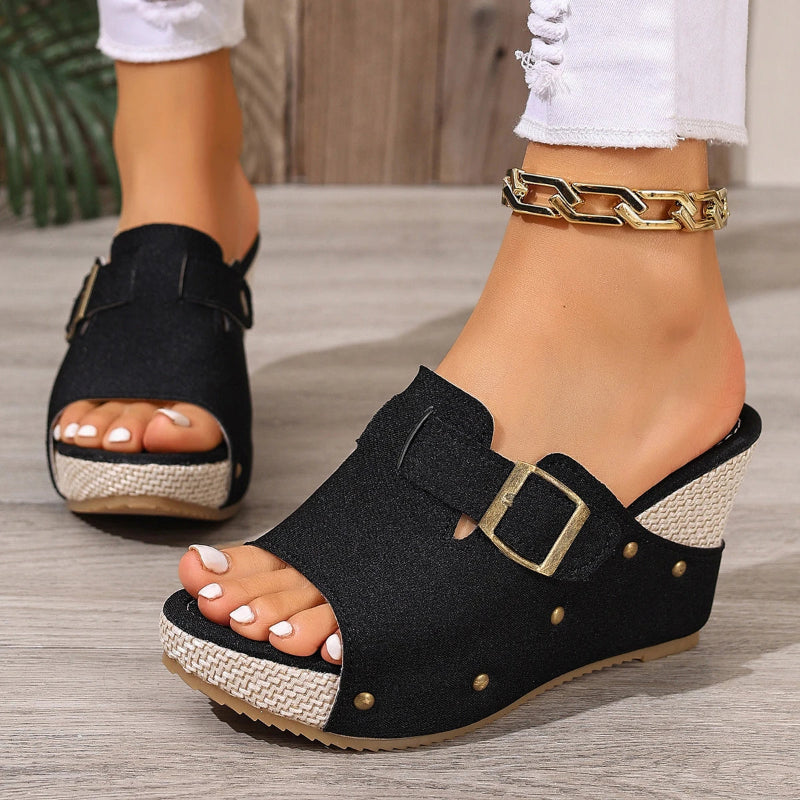 Black wedge sandals with gold buckles worn by a person on a wooden floor.