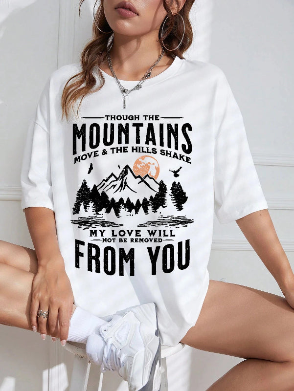 White t-shirt with mountain graphic and text worn by a person sitting on a white surface.