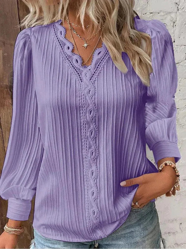 Woman wearing a purple blouse with lace details against a white brick wall.