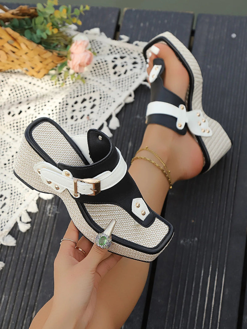 Black and white sandals with decorative straps held by a hand on a wooden surface.