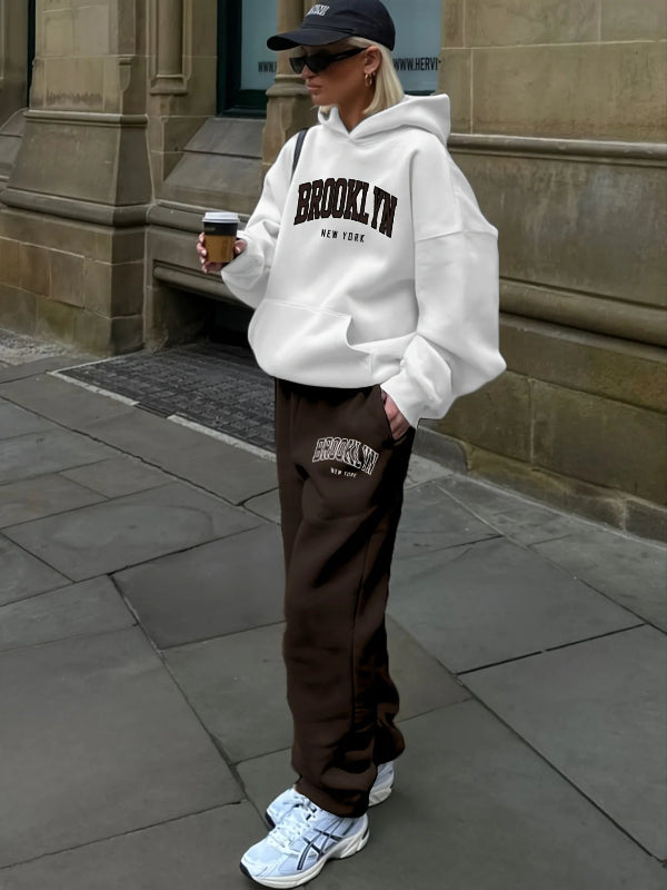 Person wearing a white 'Brooklyn New York' hoodie and brown pants, standing on a sidewalk.