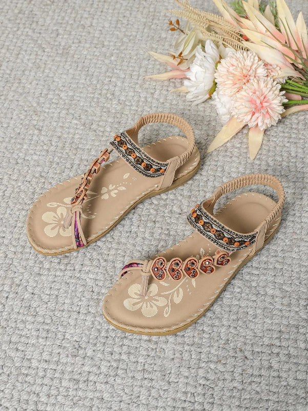 Pair of beige sandals with floral embroidery and embellishments on a textured surface with flowers.