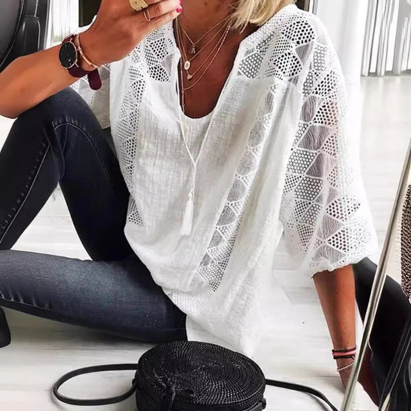 Woman wearing a white lace top and dark jeans sitting on a white surface with a black woven bag beside her.