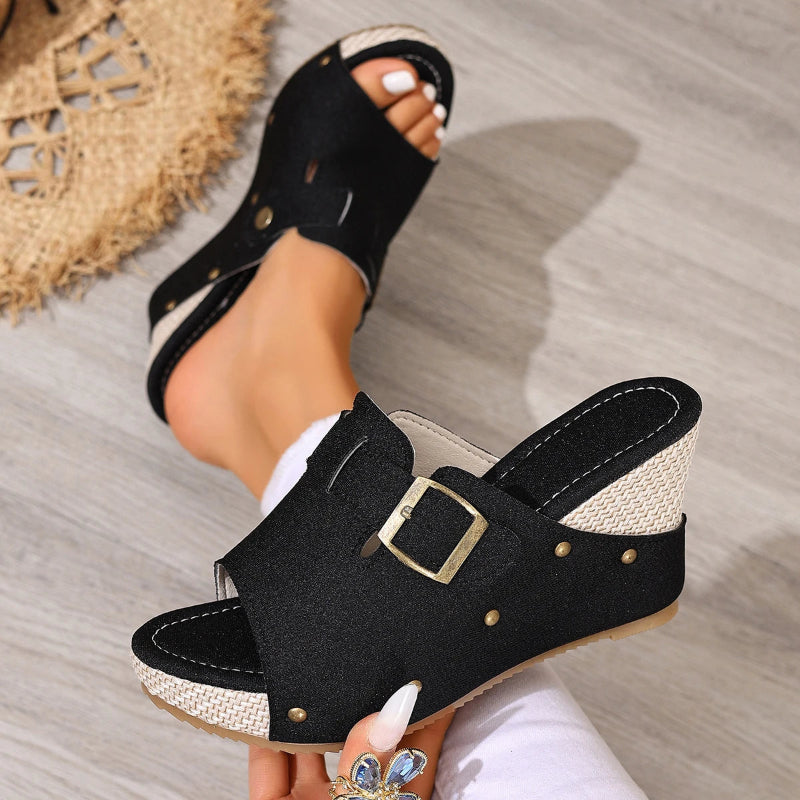 Black open-toe sandals with a gold buckle worn by a person on a wooden floor.
