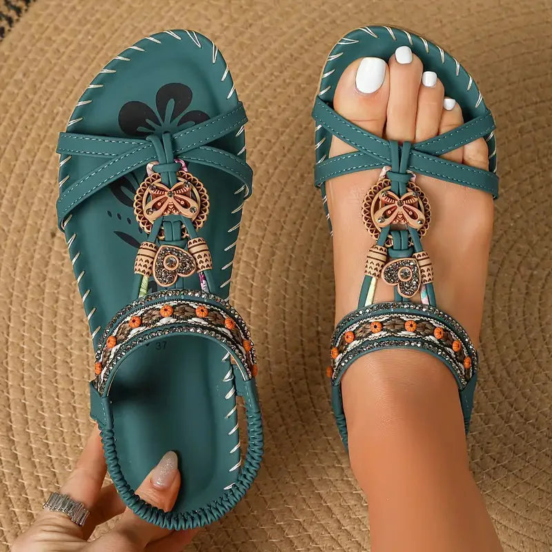 A pair of teal flat sandals with decorative woven and metallic embellishments, shown on a woven tan surface with one sandal worn on a foot and the other held by a hand.