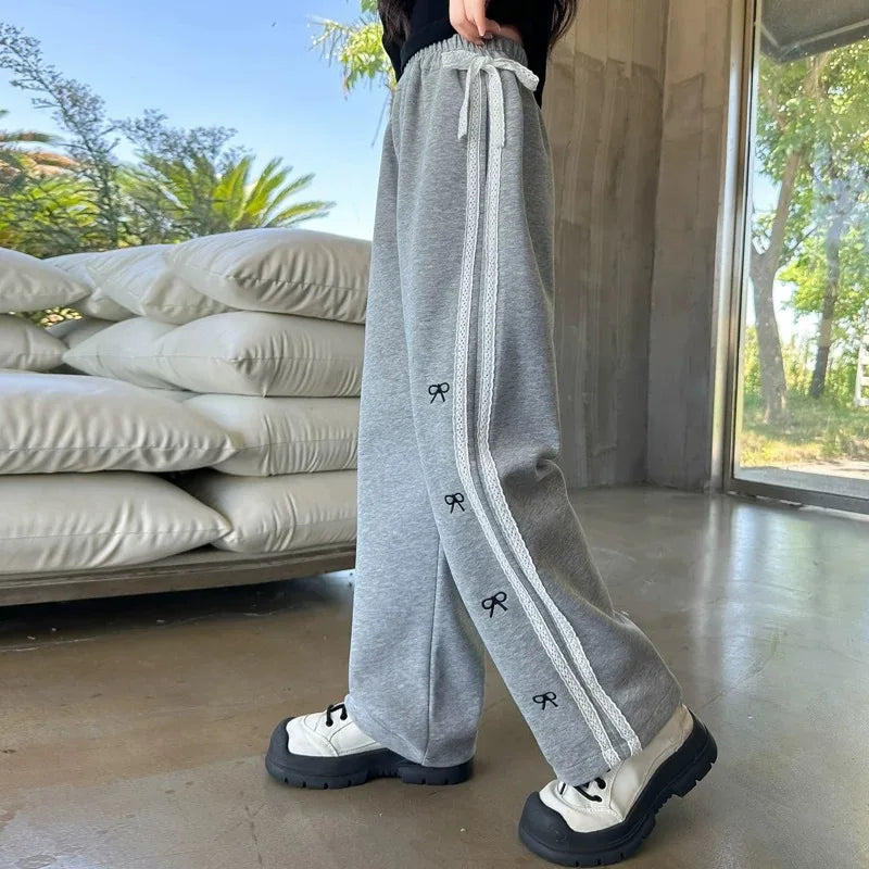 Person wearing gray sweatpants with white drawstrings and black logo details, standing in a room with large windows and outdoor view.