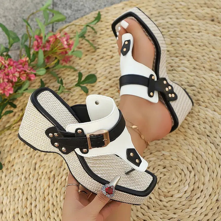 White and black sandal with decorative straps held by a hand on a woven mat with flowers in the background.
