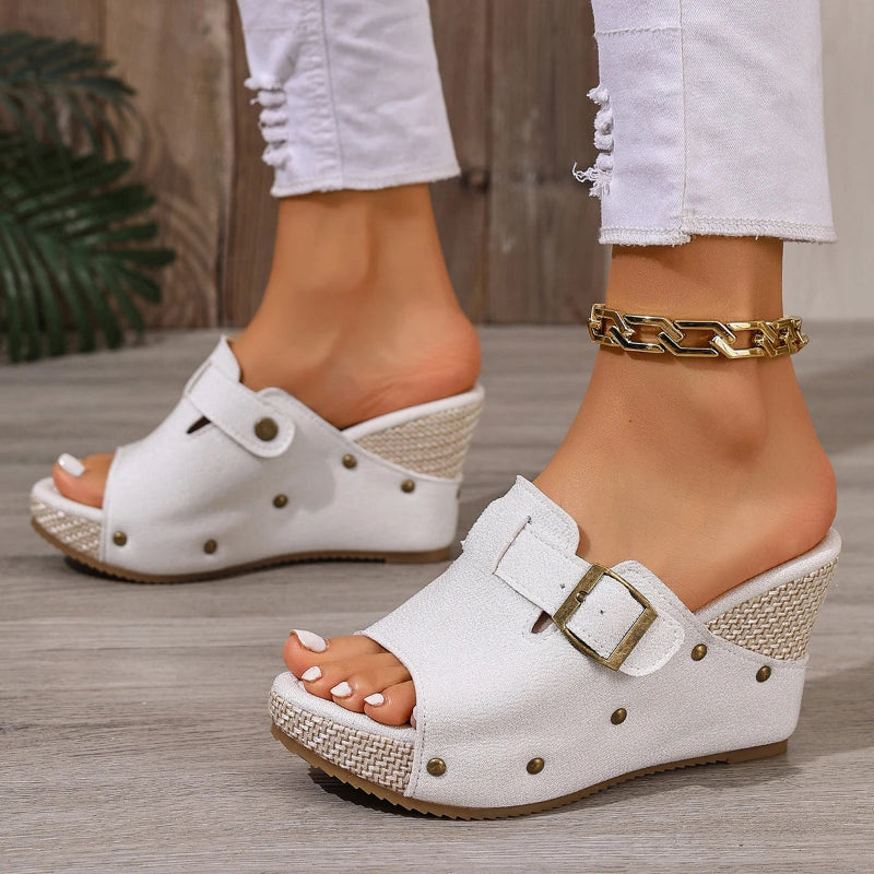 White wedge sandals with gold buckles worn by a person on a wooden floor.