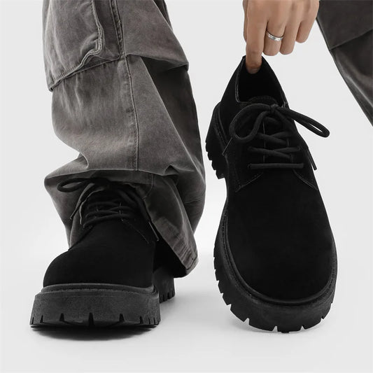 Black suede shoes with thick sole worn by a person in gray pants on a white background