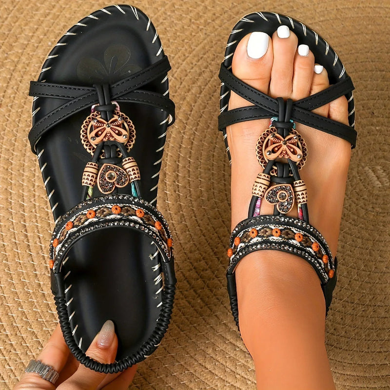 Black sandals with decorative straps and charms held by a person on a textured brown background