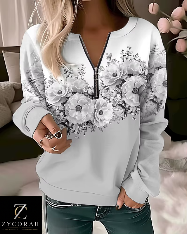 A woman wearing a white long-sleeve sweatshirt with a black floral print across the chest and a black half-zip neckline. She is also wearing dark green jeans, and a ZYCORAH brand logo watermark is visible in the bottom left corner.