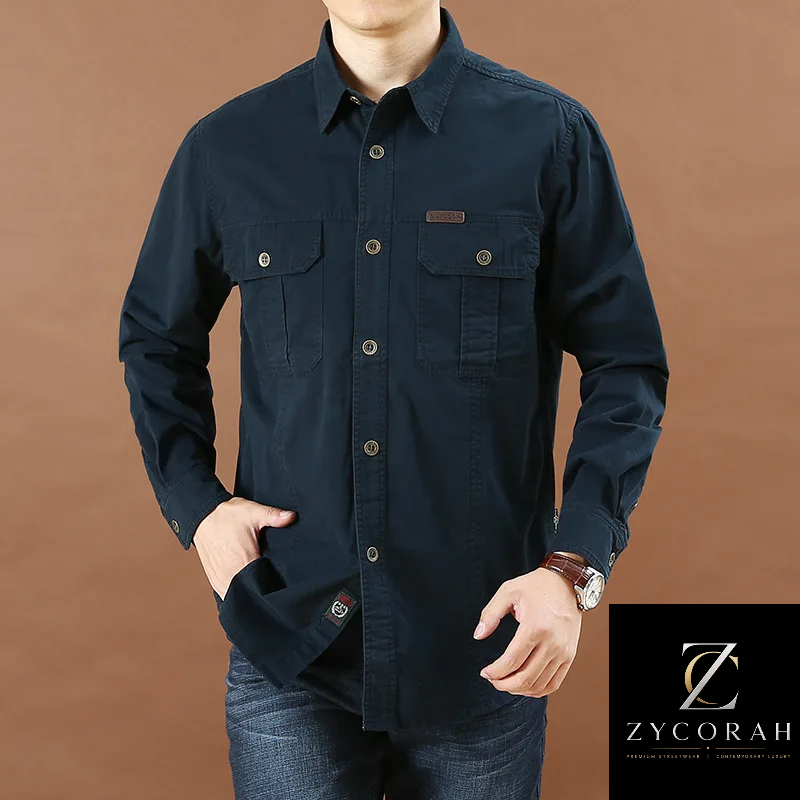 A man wearing a dark navy blue long-sleeve button-up shirt with two chest pockets, paired with blue jeans, set against a solid brown background.