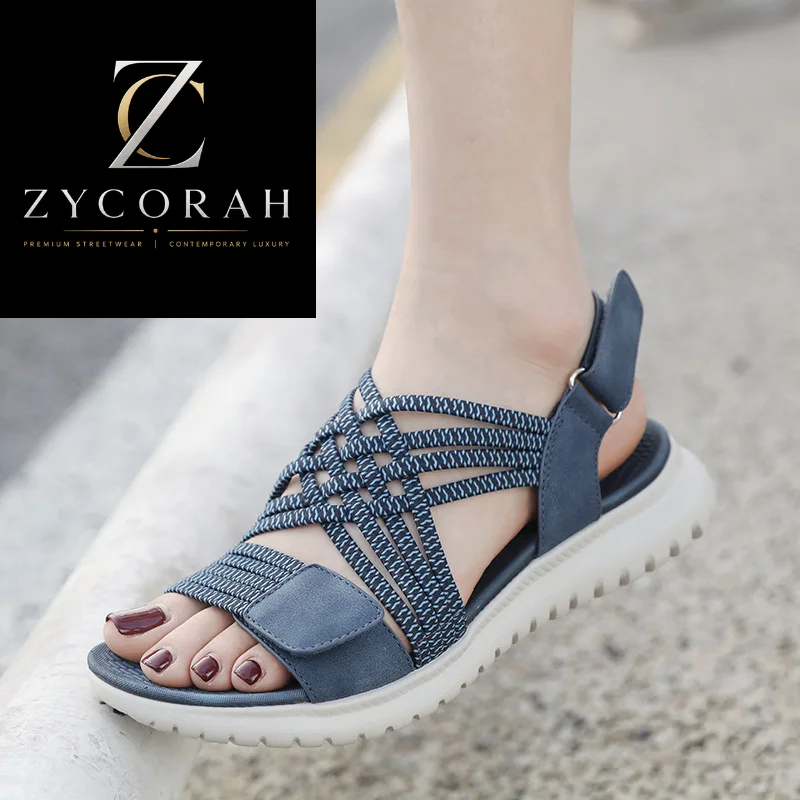 A woman's foot wearing a blue and white woven strappy sandal with a thick white sole, standing on a concrete surface. A black brand logo overlay for 'Zycorah' is in the top-left corner.