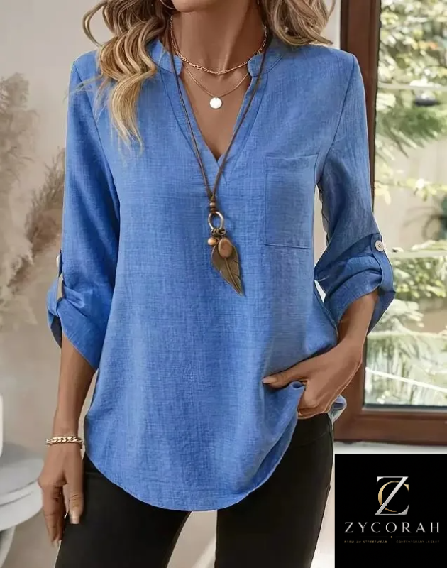 A woman wearing a blue long-sleeve linen blouse with rolled sleeves, a chest pocket, and a split neckline, paired with black pants. The Zycorah brand logo is visible in the bottom right corner.