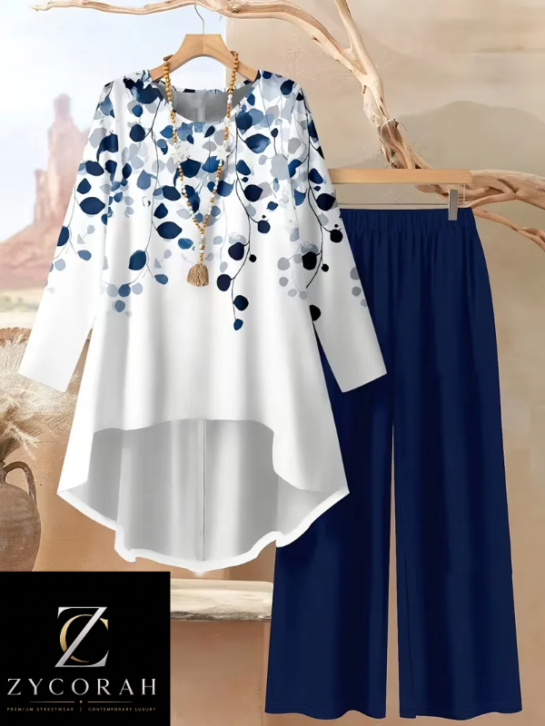 A women's two-piece outfit set featuring a white long-sleeve high-low top with a blue and navy floral vine print, paired with solid navy blue wide-leg pants. The set is displayed on wooden hangers against a warm desert landscape backdrop with a Zycorah brand logo overlay in the bottom left corner.