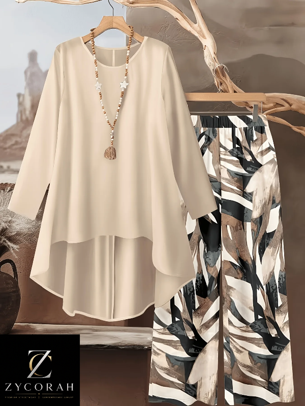A women's two-piece outfit set by ZYCORAH, featuring a cream-colored long-sleeve high-low tunic top and matching wide-leg trousers with a black, white, and brown abstract pattern. The set is styled on wooden hangers with a long beaded necklace, set against a scenic background of a desert landscape and a tree trunk.