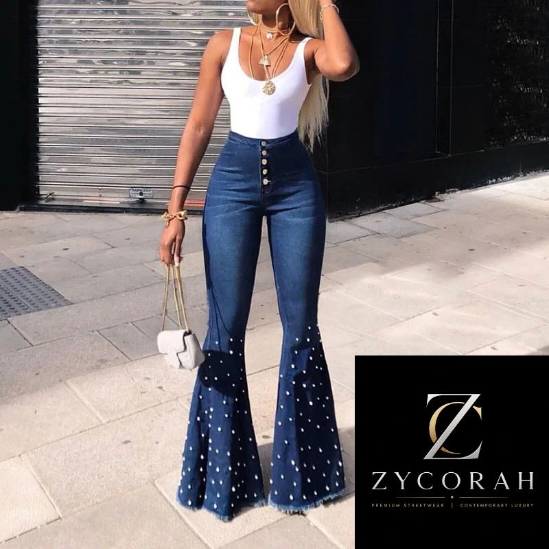 A woman wearing high-waisted blue denim flare jeans with white polka dots and button details, paired with a white sleeveless top, standing outdoors on a paved walkway. A Zycorah brand logo overlay is visible in the bottom right corner.