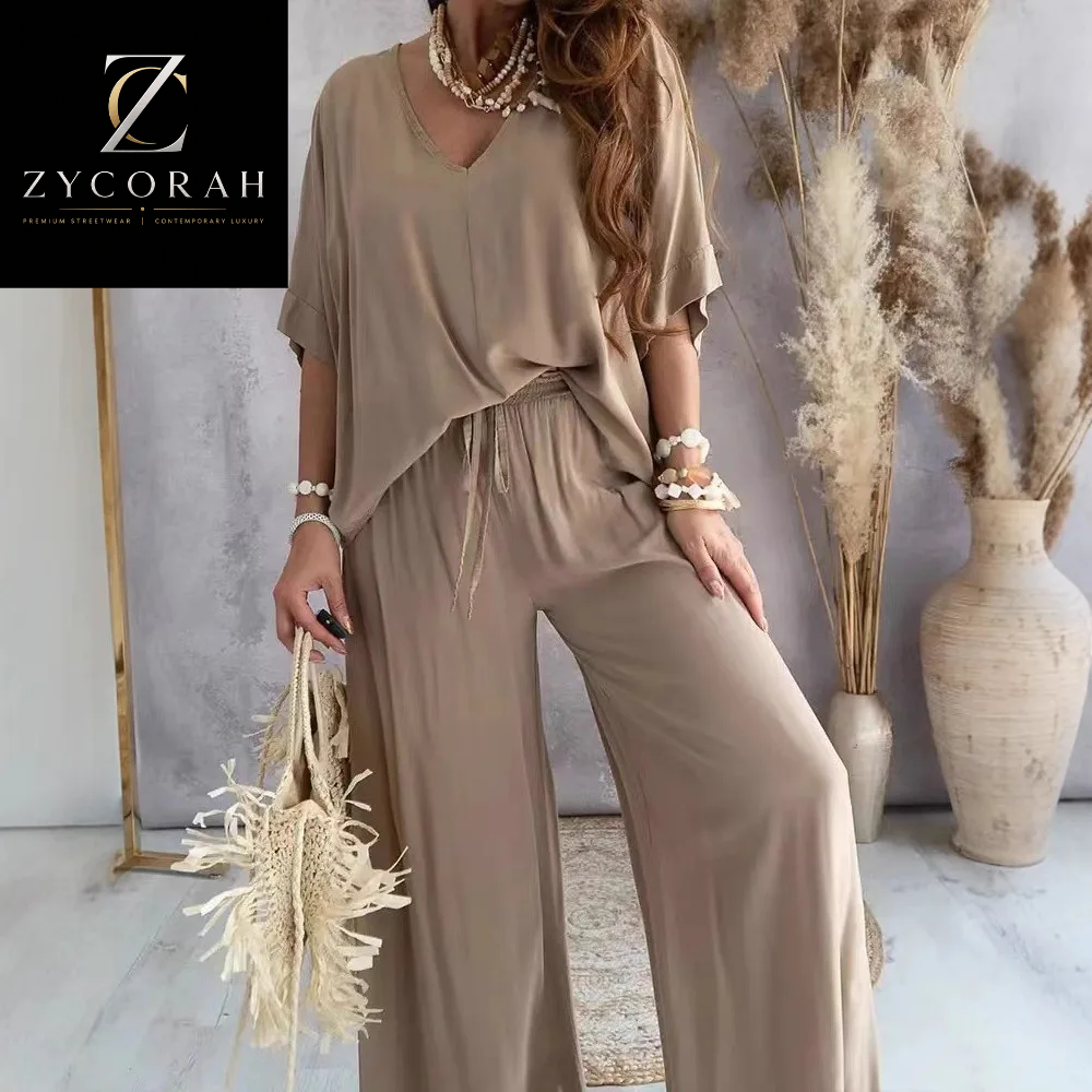 A woman models a two-piece taupe lounge set consisting of a short-sleeve V-neck top and matching wide-leg pants. The image is set in a styled interior with pampas grass in a vase and a gold-framed mirror, with a black brand overlay in the top-left corner reading 'ZYCORAH'.
