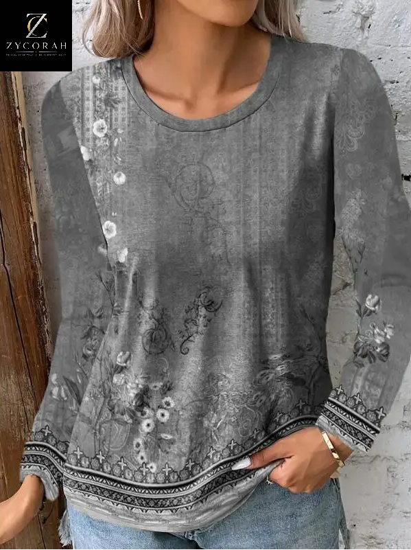 A woman wearing a gray long-sleeve top with a distressed floral and botanical print, featuring a patterned hem and cuffs, paired with light blue jeans. The Zycorah brand logo is visible in the top left corner.