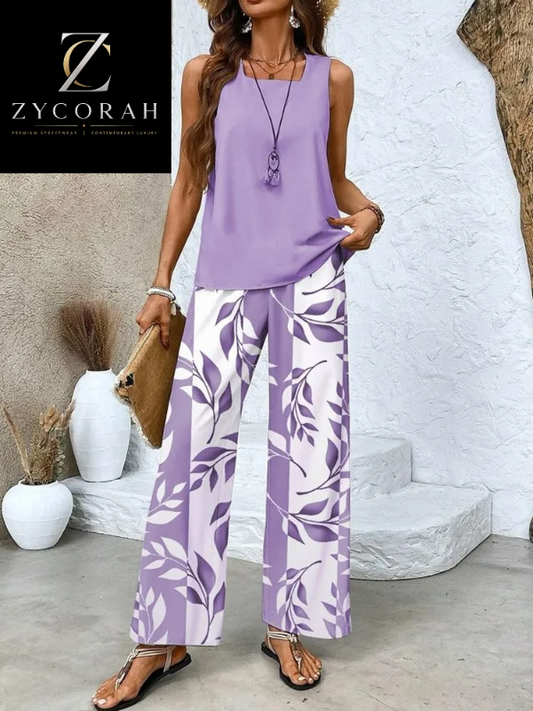 A woman models a two-piece lavender and white leaf-patterned set consisting of a sleeveless top and wide-leg pants, styled with a brown clutch and sandals in an outdoor patio setting.