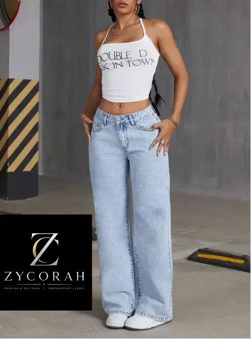 A model wearing a white halter crop top with 'DOUBLE D SKIN IN TOWN' text and light-wash wide-leg jeans, standing in a gray industrial setting. A Zycorah brand logo overlay is visible in the bottom left corner.