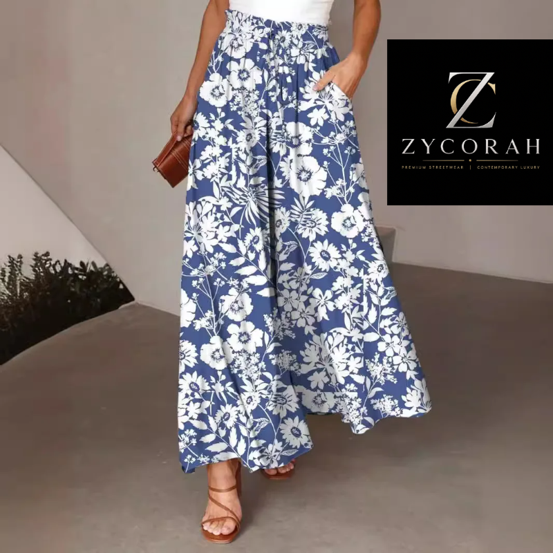 A model wears high-waisted, wide-leg maxi pants featuring a blue and white floral pattern, styled with a white top and brown strappy sandals. A black brand logo overlay for 'Zycorah' is visible in the upper right corner.