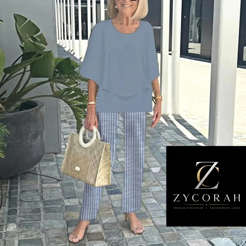 A woman stands barefoot on a stone walkway wearing a light blue short-sleeve top and blue-and-white striped pants, holding a woven beige handbag. A Zycorah logo overlay appears in the bottom right corner.