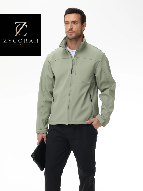 A man wearing a light sage green Zycorah softshell jacket over a white t-shirt and black pants, holding a black folder against a white studio background with a Zycorah brand logo in the top left corner.