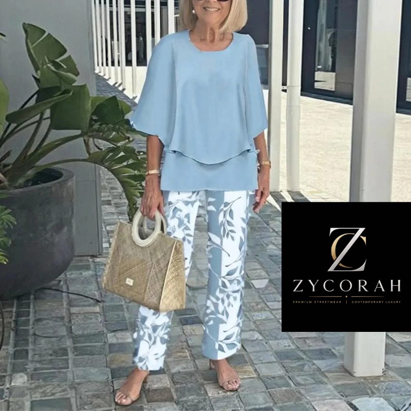An older woman stands outdoors wearing a light blue short-sleeve top and white pants with a blue leaf pattern, holding a woven tan handbag. A black Zycorah logo overlay appears on the right side of the image.