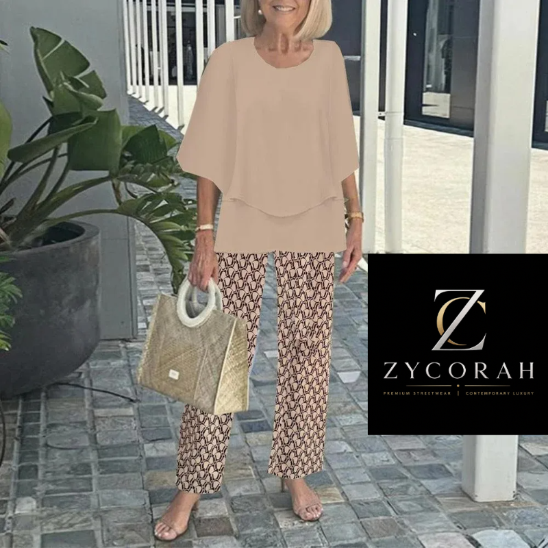 A woman stands barefoot on a stone walkway wearing a beige short-sleeve draped top and brown-and-white patterned pants, holding a woven beige handbag. A Zycorah logo overlay appears in the bottom right corner.