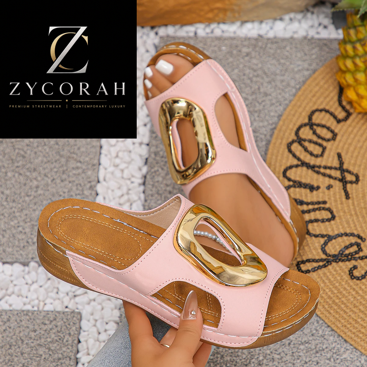A pair of women's slide sandals in light pink faux leather with gold-tone metal accents, shown being worn and held by a hand. The brand logo 'ZYCORAH' is displayed in a black box in the top-left corner.