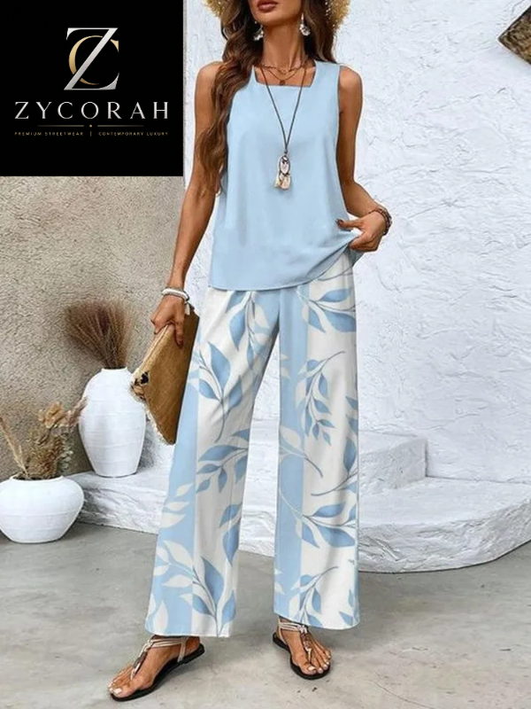 A woman models a sleeveless light blue top paired with wide-leg pants featuring a light blue and white leaf pattern. She is standing in a styled indoor setting with a textured white wall, decorative vases, and a black brand logo overlay for ZYCORAH in the top-left corner.