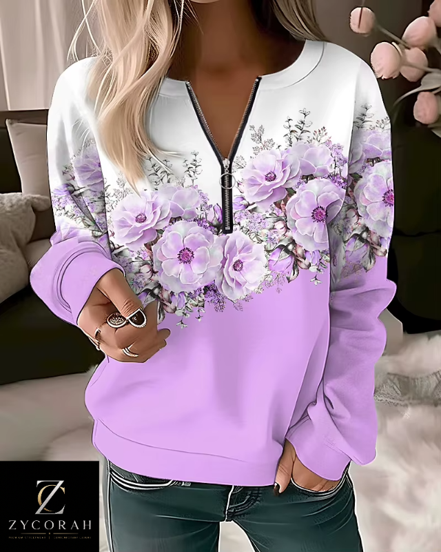 A woman wearing a lavender and white long-sleeve sweatshirt with a floral print across the chest and shoulders, paired with dark green jeans. The sweatshirt features a half-zip neckline with a black zipper.