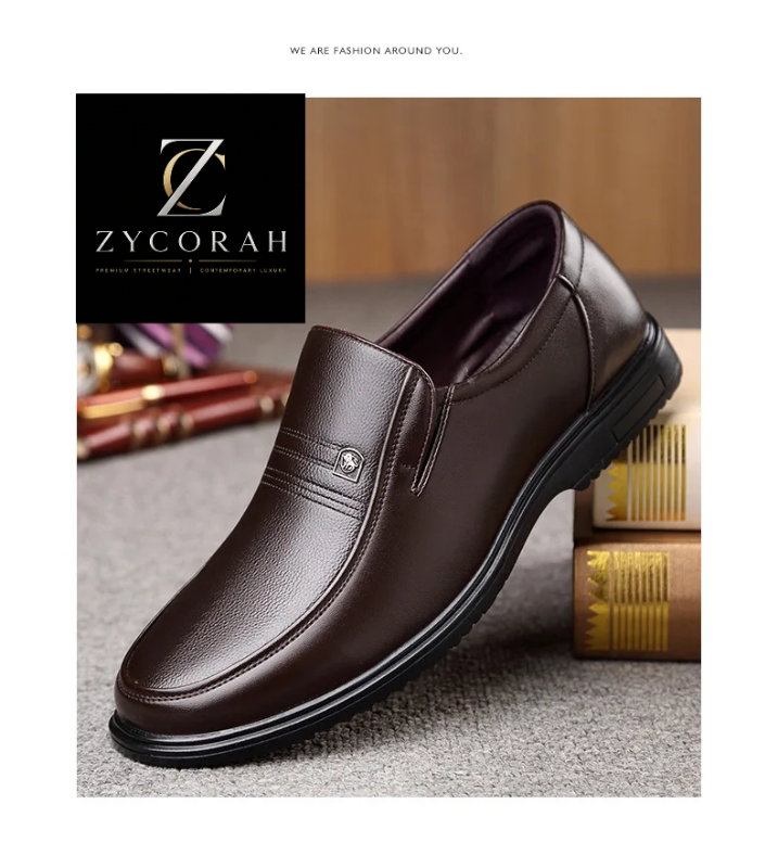 A dark brown leather slip-on loafer for men, styled on a textured gray carpet next to stacked books. The shoe features a decorative metal emblem on the side and a black sole.
