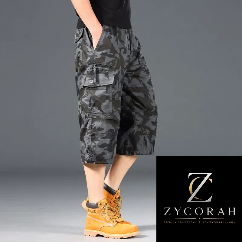 A model wearing ZYCORAH brand dark gray and black camouflage cargo shorts, paired with a black t-shirt and yellow boots, against a gray studio background with a brand logo overlay in the bottom right corner.