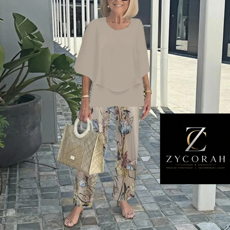 An older woman stands barefoot on a stone walkway outdoors, wearing a beige short-sleeve draped top and beige wide-leg pants with a blue and floral print. She holds a woven beige handbag.