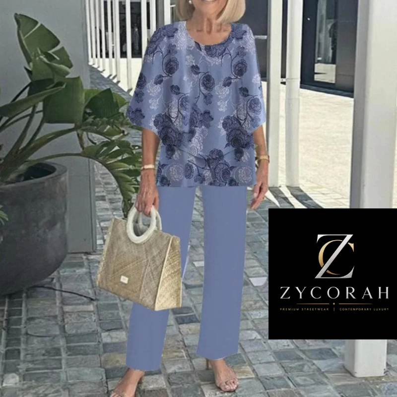 A woman wearing a light blue and navy floral two-piece set with wide sleeves and straight-leg pants, holding a woven beige handbag. The image is set on a stone walkway with a modern building background, and features a black brand logo overlay for ZYCORAH in the bottom right corner.