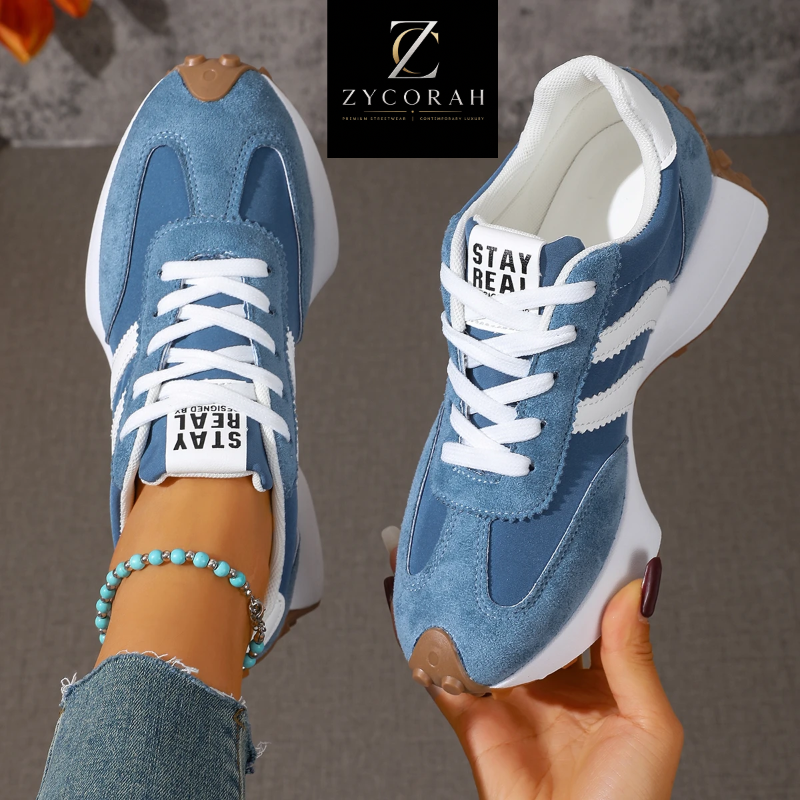 A pair of blue and white chunky sneakers shown in a lifestyle presentation, with one shoe worn on a foot and the other held in a hand. The brand logo 'Z YCORAH' is displayed at the top, and 'STAY REAL' is printed on the tongue labels.