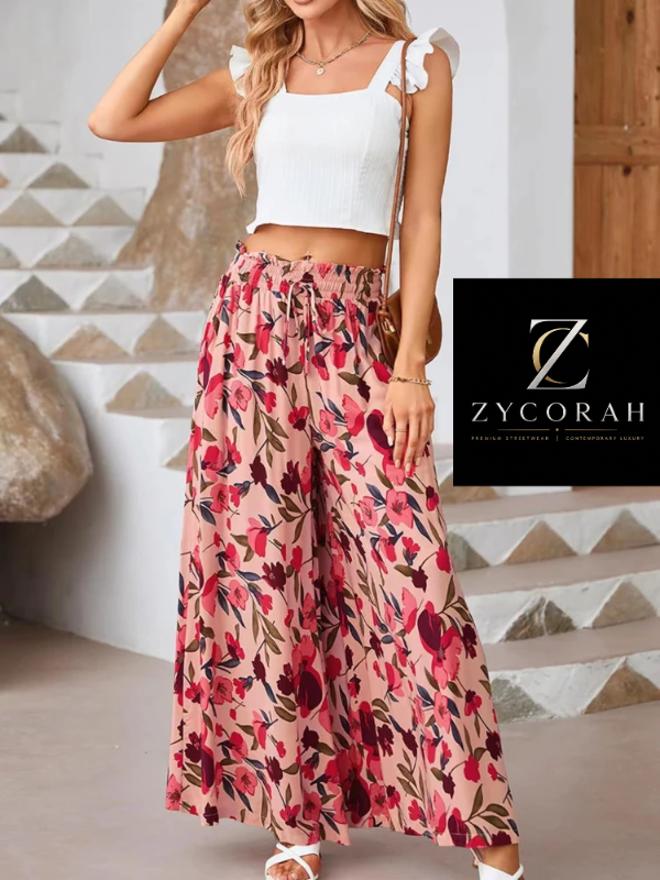 A woman models a white ribbed crop top with ruffled shoulder straps and high-waisted, wide-leg floral pants in pink, red, and dark brown tones. She is standing in front of a modern geometric stone staircase.