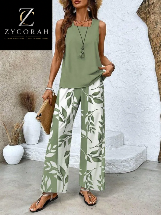 A woman models a sleeveless sage green top paired with wide-leg pants featuring a green leaf pattern on a white background. She is holding a brown clutch and wearing brown sandals, set against a rustic white wall backdrop with decorative vases.