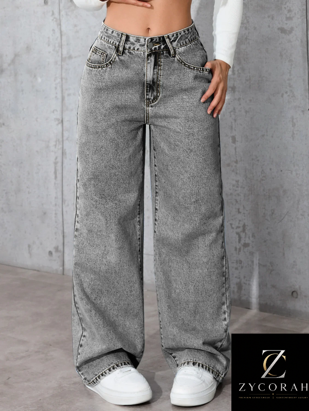 A person wearing wide-leg gray denim jeans with a faded wash, paired with a white long-sleeve top and white sneakers, standing against a textured gray concrete wall. A Zycorah logo is visible in the bottom right corner.