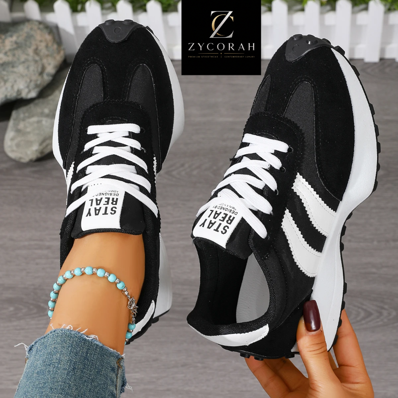 A pair of black and white low-top sneakers shown in a lifestyle setting, with one shoe worn on a foot with a beaded ankle bracelet and the other held by a hand. A black brand label for 'ZYCORAH' is displayed at the top center.
