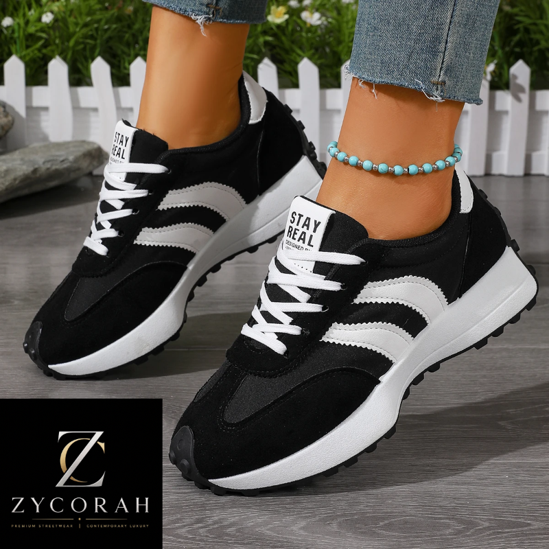 A pair of black and white low-top sneakers with white laces and textured panels, shown on a model's feet. The shoes feature a chunky white sole and a black-and-white side stripe design.